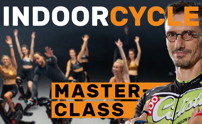 Trainer HaJü als Instructor für eine Indoor Cycle Masterclass. Im Hintergrund sind leicht unscharf die Kursteilnehmer zu sehen, sie winken in die Kamera